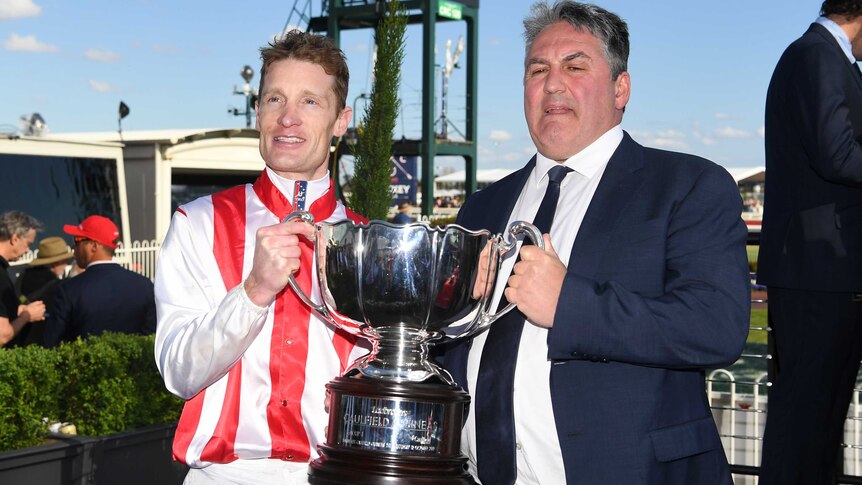 A jockey and trainer stand smiling holding a cup after winning a big race.