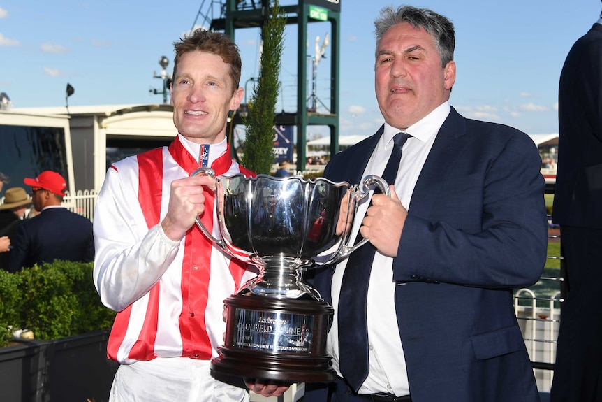 A jockey and trainer stand smiling holding a cup after winning a big race.