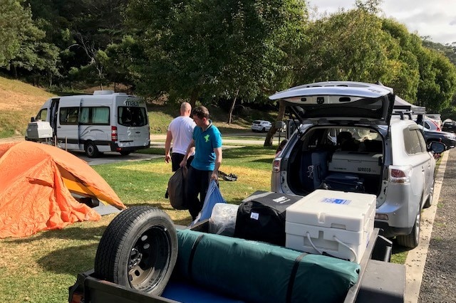 Two men walk around a vehicle with a trailer on the back as they pack up a camp site.