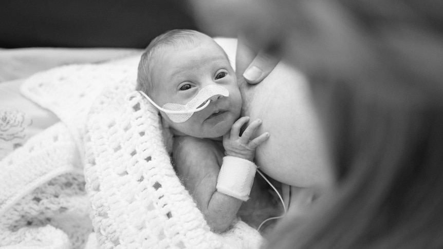 A pre-term baby with a nasal gastric tube laying on her mother's chest , tucked up in a knitted blanket.