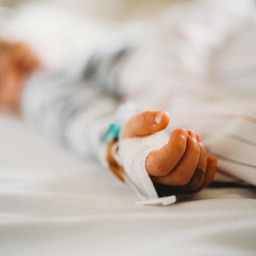 In focus is the hand of a child laying in a hospital bed with the background blurred.