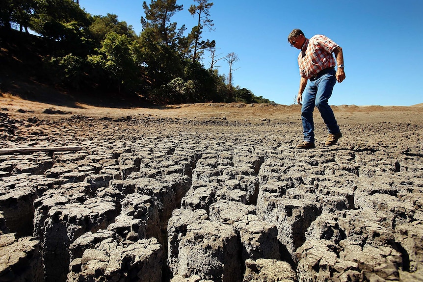 A man wearing jeans and a checked shirt walks along a dried lake bed.