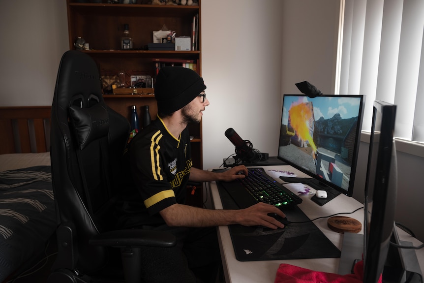 A teenager wearing a beanie sits at a desk in his bedroom, playing a computer game on one of two monitors.