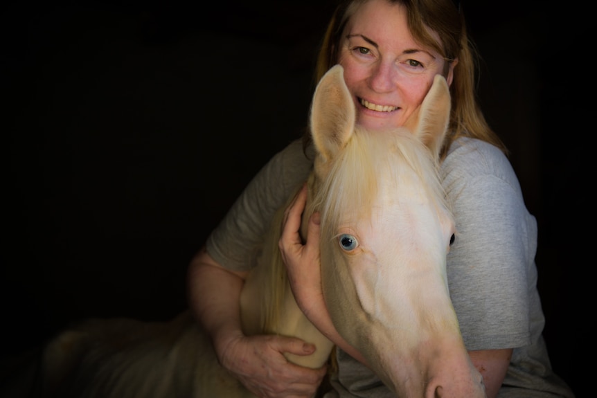 A portrait of Jill Barton smiling and cuddling a small white horse.