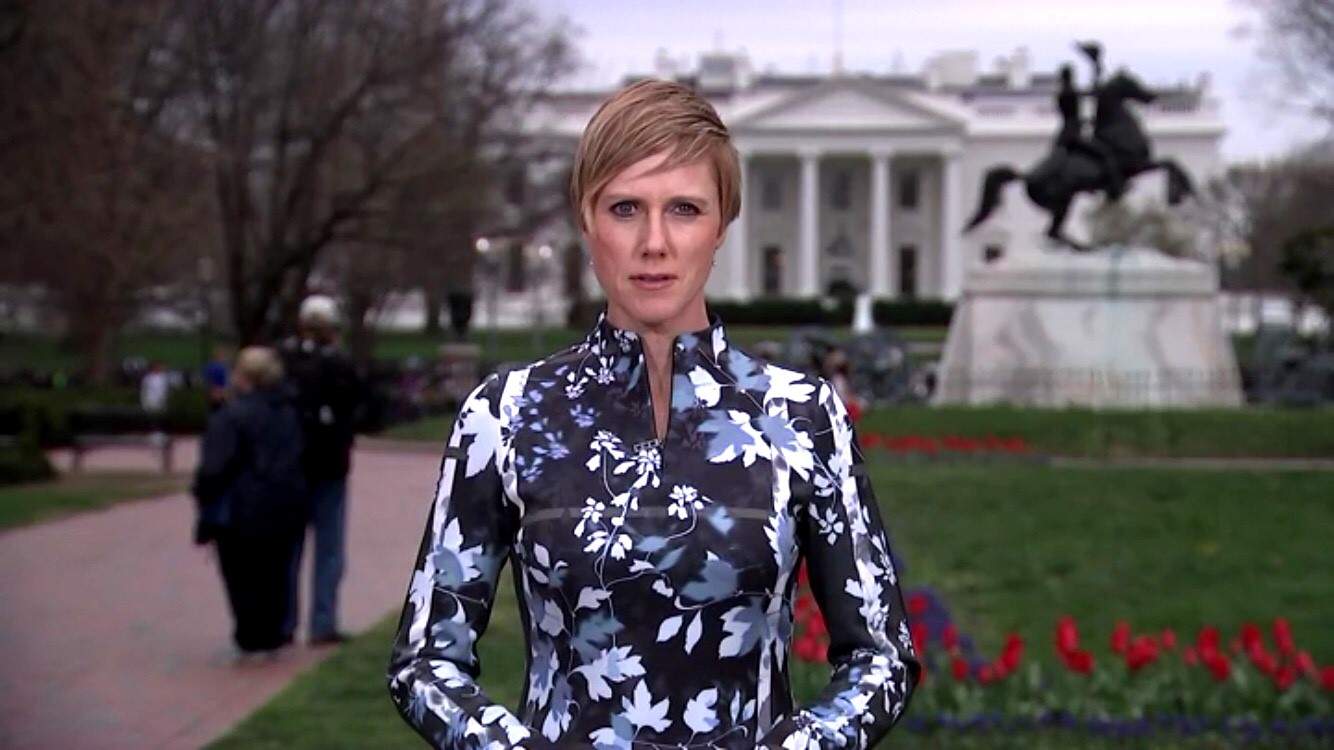 A news reporter stands some distance from the White House in Washington looking serious.