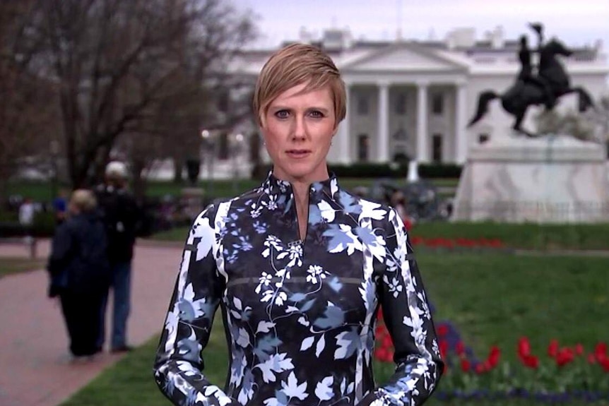 A news reporter stands some distance from the White House in Washington looking serious.