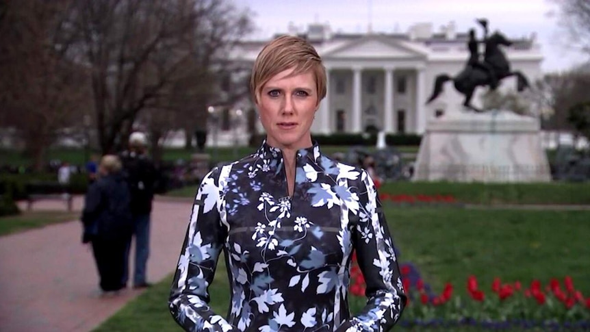 A news reporter stands some distance from the White House in Washington looking serious.