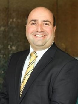 A smiling man in a dark suit and yellow ties smiles at the camera 