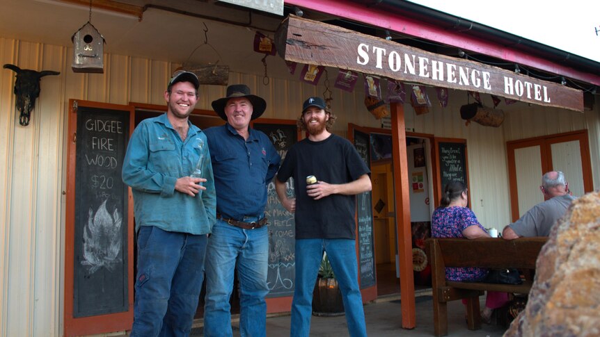 Three men stand outside of a small, old pub smiling. A sign reads 'Stonehenge Hotel'.