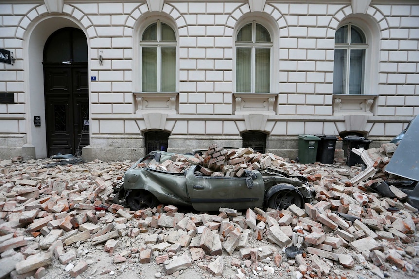 You see a small green hatchback crushed under a pile of bricks in front of a white neoclassical building.