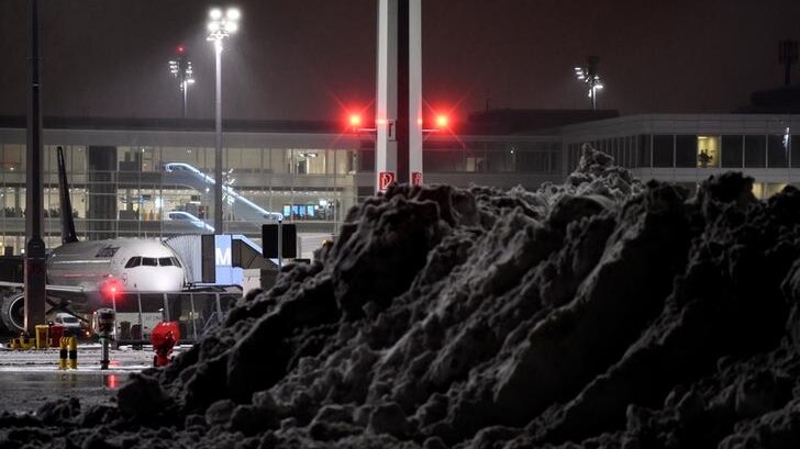 A mound of snow stands in the foreground with a plane in the background and airport behind it in the dark.