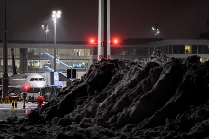 A mound of snow stands in the foreground with a plane in the background and airport behind it in the dark.