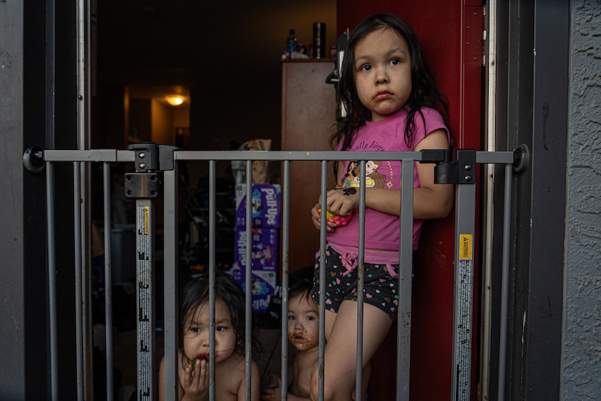 Three young girls stand behind a baby gate at the door to a motel room.