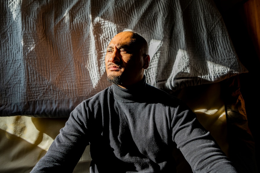 A man sits against a mattress. He is looking into the light from a nearby window.