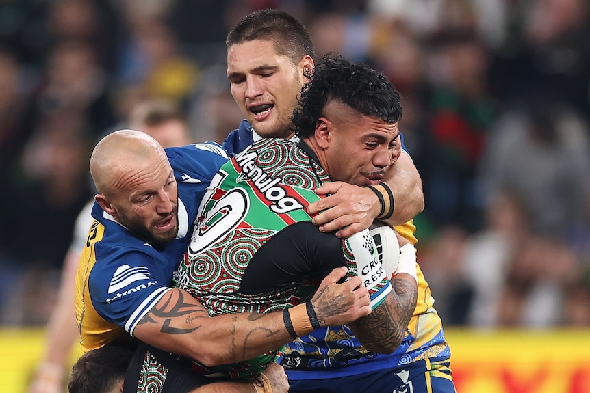 A South Sydney NRL is tackled by two Parramatta opponents while holding the ball.