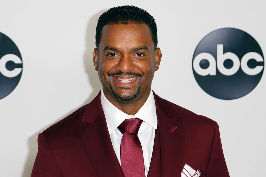 Alfonso Ribeiro wears a dark red suit with a white and red lined pocket square and red tie. He has a wide smile on his face.