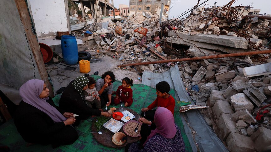 Three women and four children eat off a plate amid rubble in Rafah.