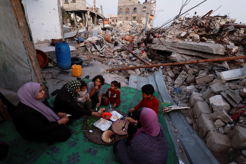 Three women and four children eat off a plate amid rubble in Rafah.