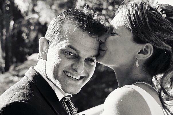 Black and white photo of a bride and groom where she is kissing him on the side of the head, he smiles at the camera.