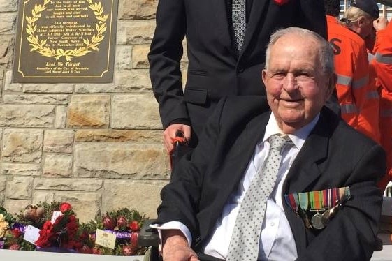 elderly man in suit with war medals, smiling, sitting in wheel chair. young man in black suit standing behind him