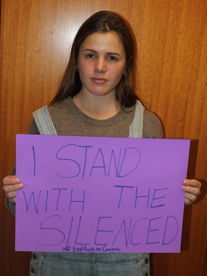 Girl stands with purple sign reading: I stand with the silenced #EndRapeonCampus