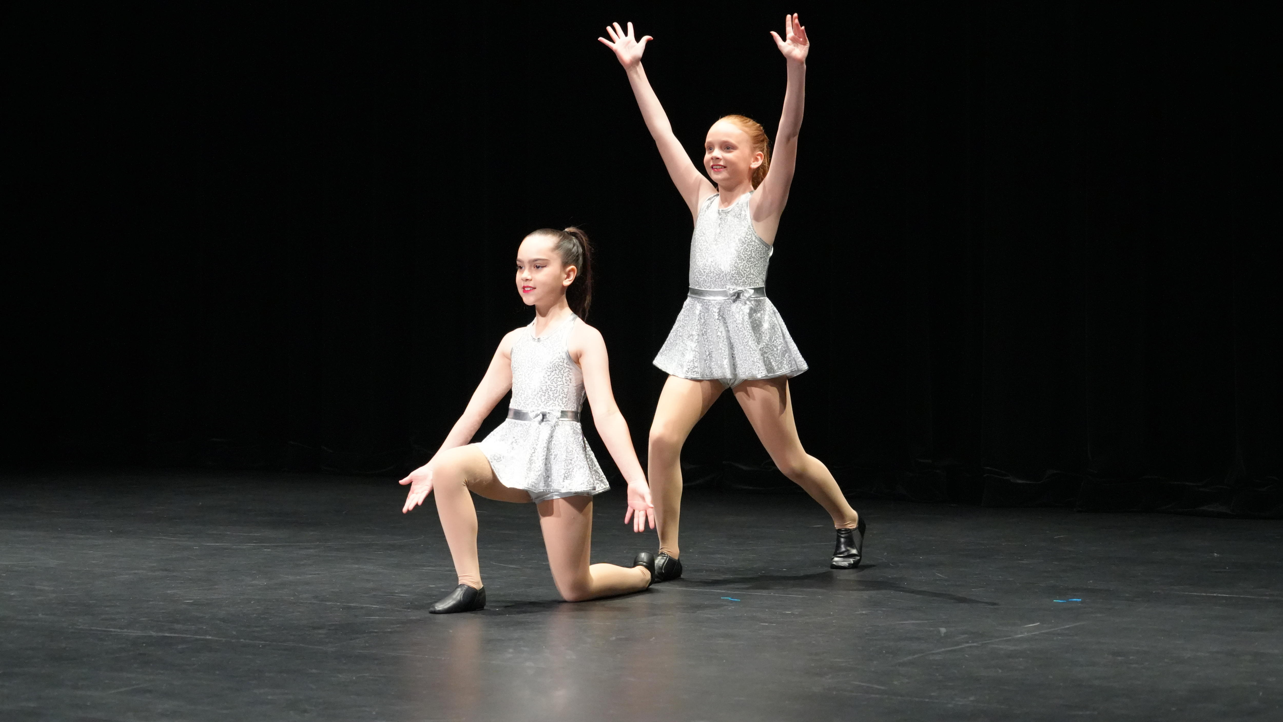 Dos niñas con trajes blancos de jazz posan frente a un escenario negro. Una tiene las manos una detrás de la otra.
