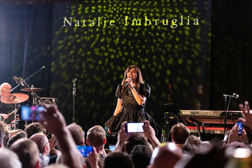 A crowd of people watch and video Natalie Imbruglia sing on stage, her name is projected onto a wall behind her