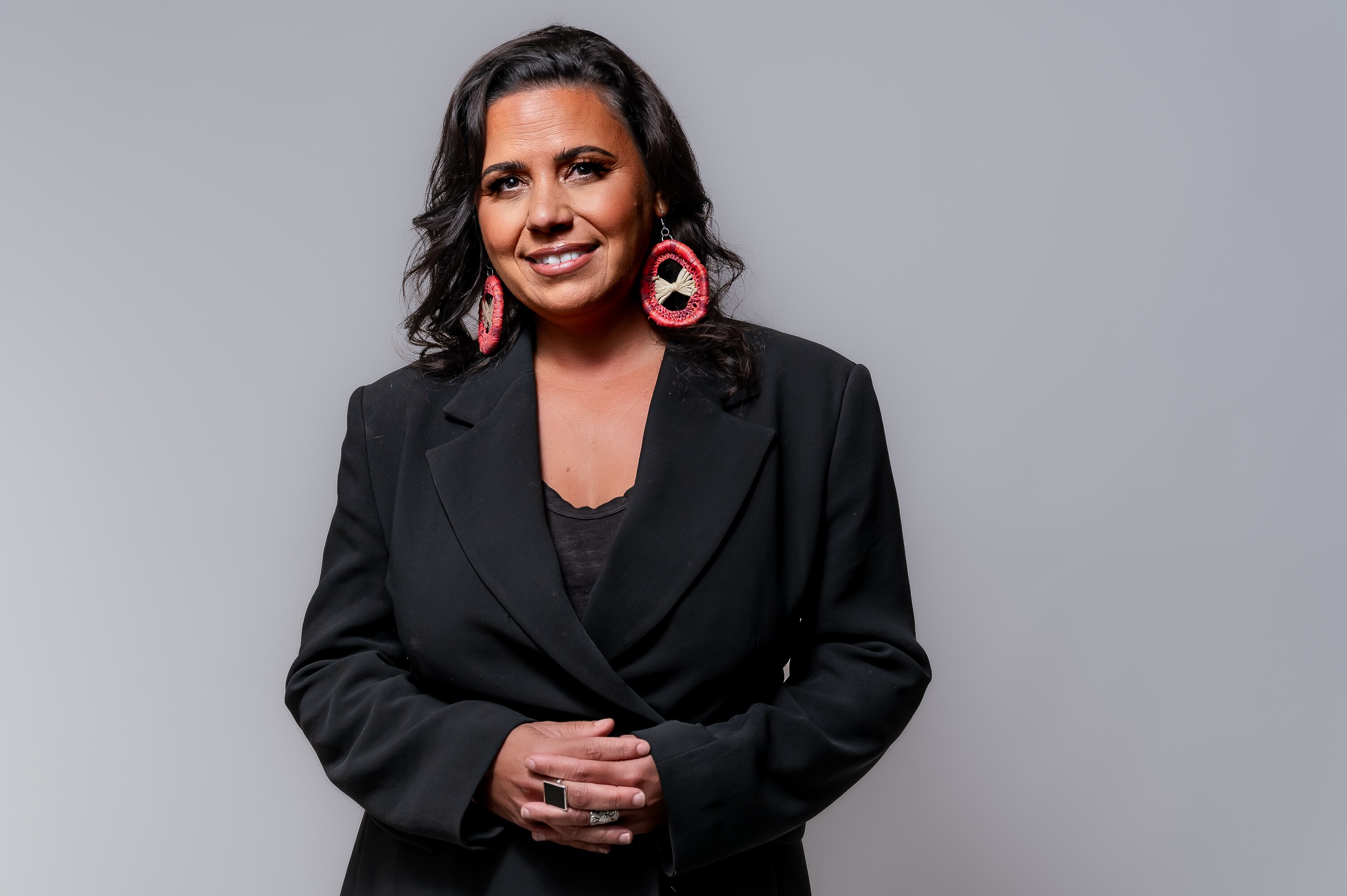 Smiling dark haired woman with bright weaved earrings in black top and blazer standing in front of light grey studio backdrop