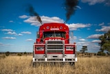 A close shot of the front of a red Mack haulage truck. Two diesel exhaust pipes are blowing black smoke. Blue sky surrounds.