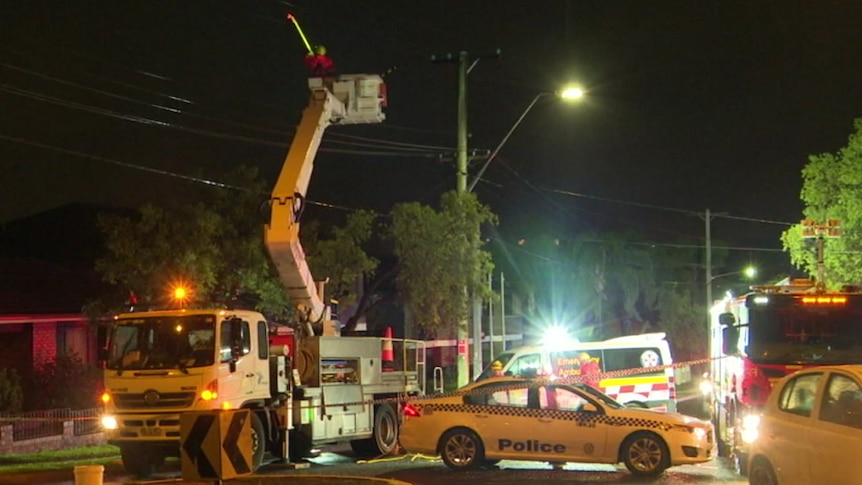 Police and ambulance park across a road while a man in a cherry picker tests electricity wires above.