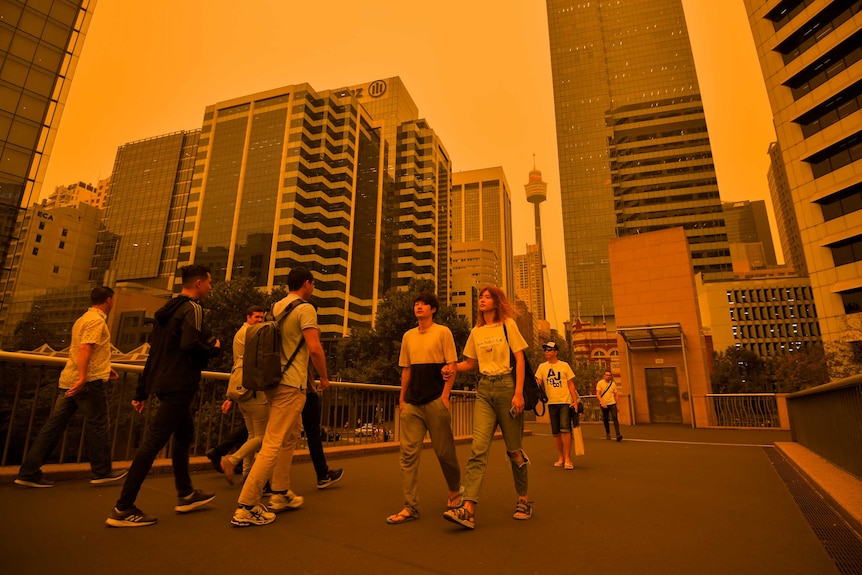 People walk across a bridge in the Sydney CBD, the sky is orange and hazy with Sydney tower in the background.