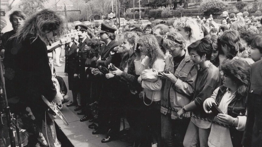 A big-haired '80s rock band stands on a train performing to a largely female crowd standing on the platform.