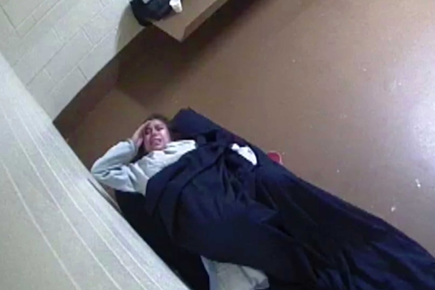 A grainy still shows a woman, mostly covered by a blanket, grimacing while laying on a small bed in an empty cell.