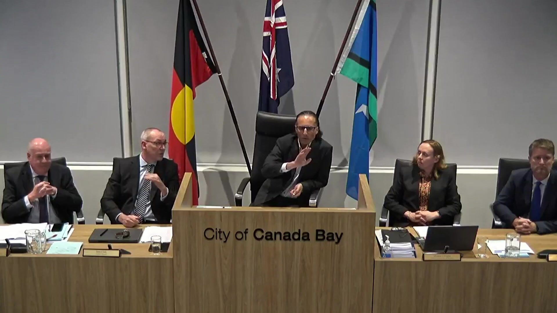 Five people sit behind a long desk at the City of Canada Bay Council with the acting mayor raising his hand while he speaks