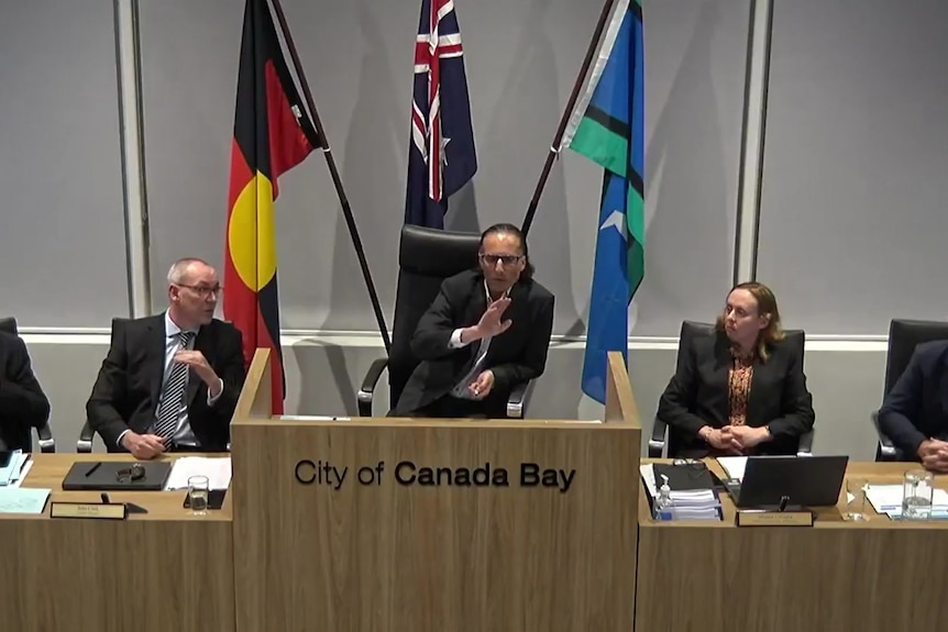 Five people sit behind a long desk at the City of Canada Bay Council with the acting mayor raising his hand while he speaks