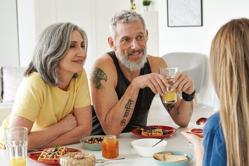 An older couple look expectantly at a young woman over breakfast. 