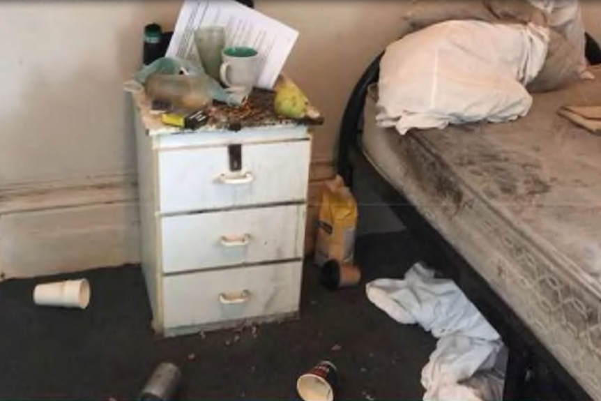 Empty cups and cigarette butts strew the floor of a bedroom, with a bare dirty mattress and a bedside table covered with filth.