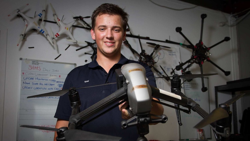 A young man Sam Hadley pictured holding a damaged drone, which other damaged drones on the wall behind him.