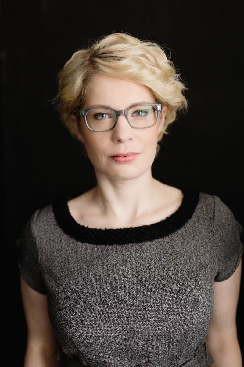 A woman with blonde, short, wavy hair and glasses in front of a black background