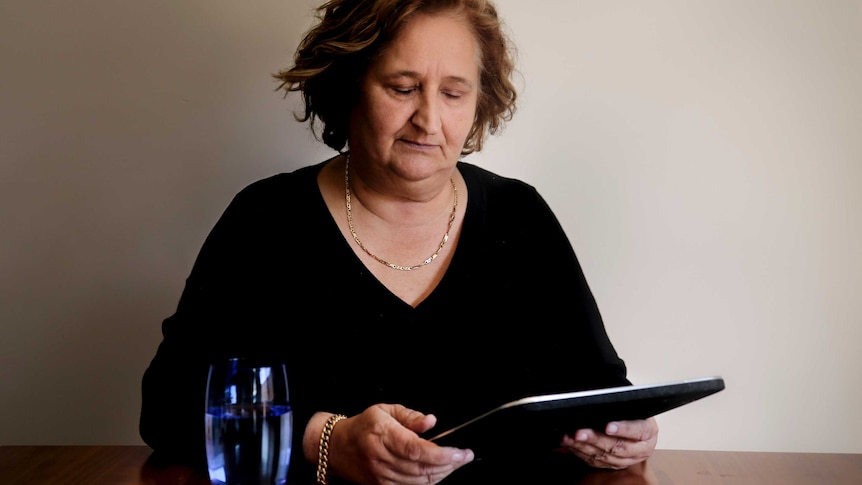 Woman looks at picture frame in hand sitting at wooden table with glass of water in front of her.