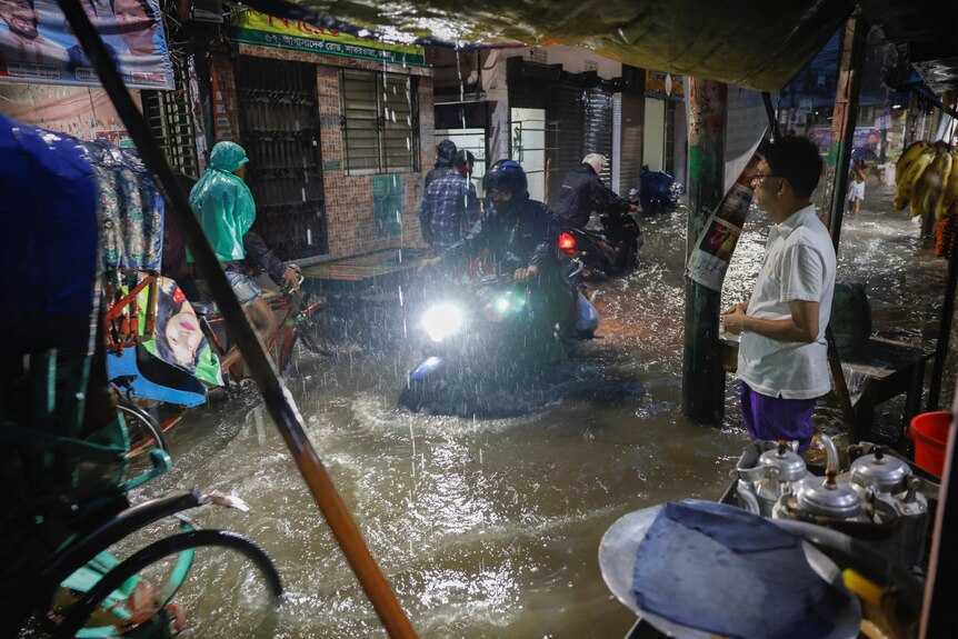 Two people ride motorbikes through knee deep water on a street as a man watches. 