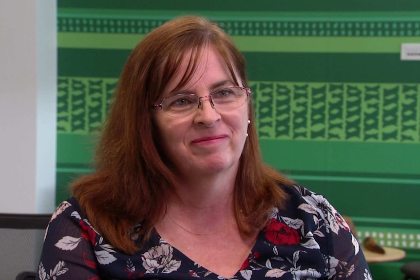 Professor Hemsley has long brown-red hair and glasses and is looking right of frame. She's sitting in front of a green wall.