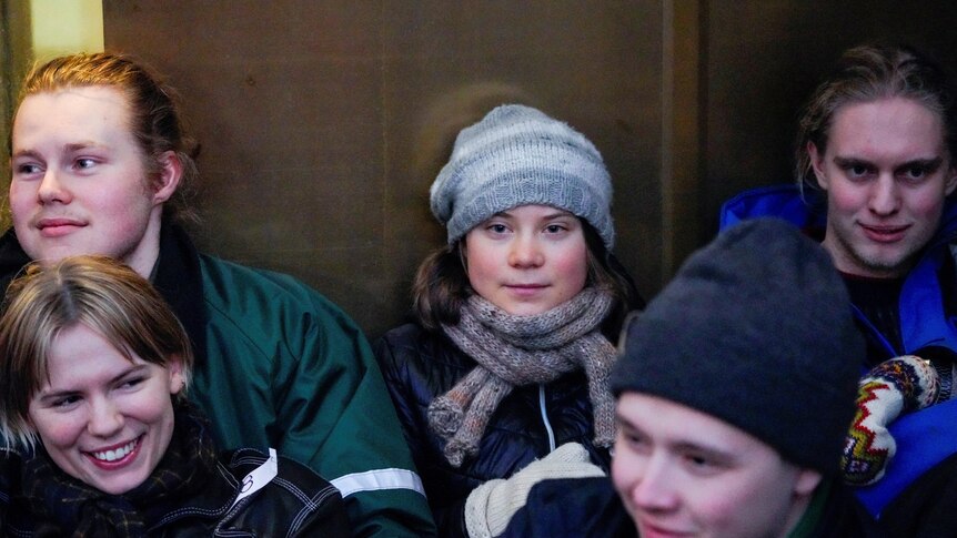 Greta Thunberg wears a beanie, scarf and jacket and is sitting on the ground next to other young people. 