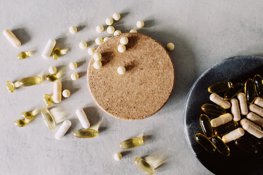 Vitamins and tablets spilled over a white table top with the lid of a container and a bowl full of pills.