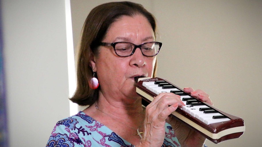 A woman wearing glasses and a floral dress plays a melodica