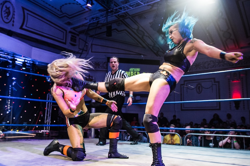 Female wrestler with blue hair air-kicking another wrestler in the ring.