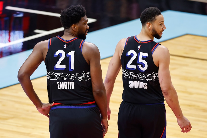 Ben Simmons and Joel Embiid look over their right shoulders wearing black singlets