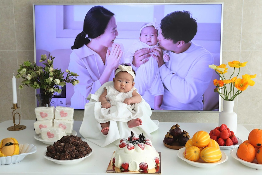 A baby girl sits on a table in white with fruits and a strawberry cake around her. A picture of her parents in the background.