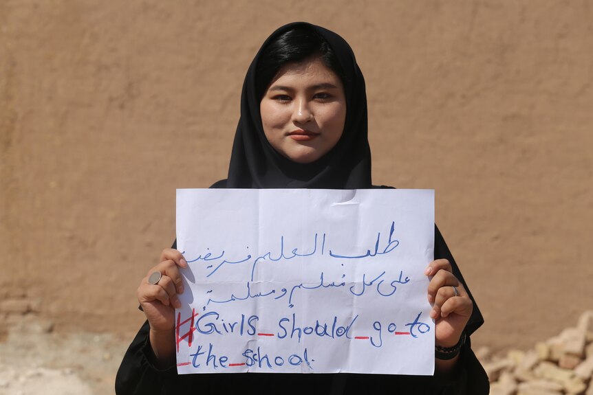 A woman in a headscarf holding a handwritten paper sign that reads 'Girls should go to school'
