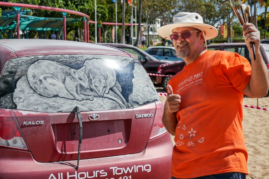 Artist Scott Wade holds paint brushes in his hand in front of an artwork painted in dirt on a car window